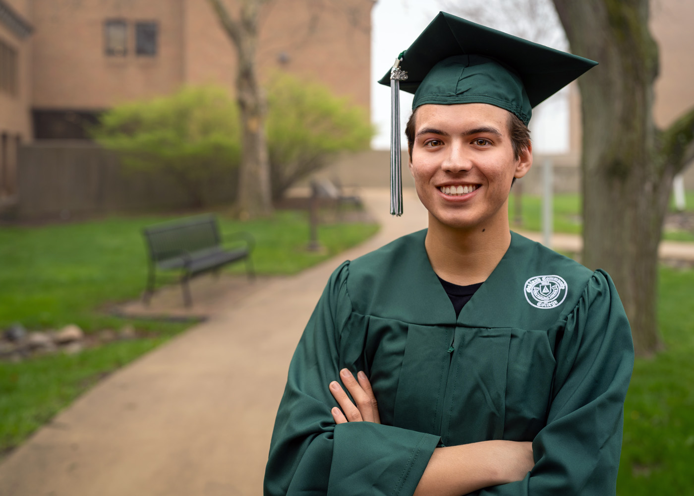 Cosmo Bray posing outside of campus. He's smiling with his arms crossed. He's wearing his green O C C cap and gown