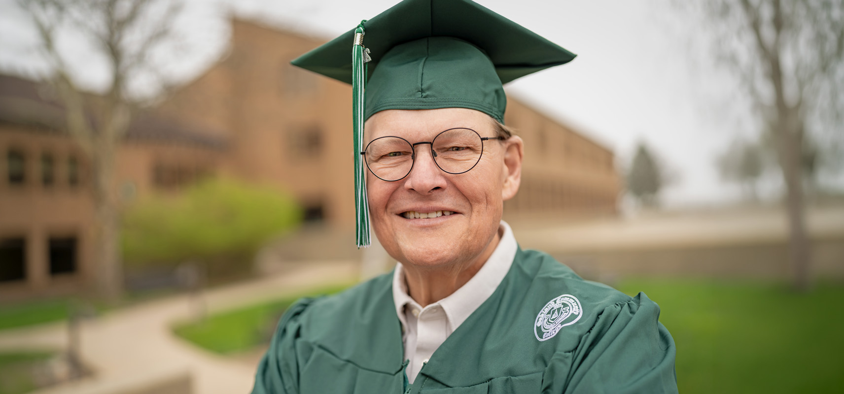 Don Fenton posing outdoors on campus. It's a close up photo and he's wearing his green o c c graduation cap and gown