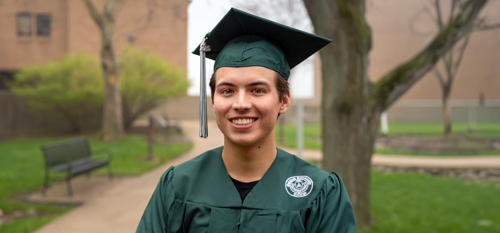 Cosmo Bray posing outdoors on campus. It's a close up photo and he's wearing his green o c c graduation cap and gown