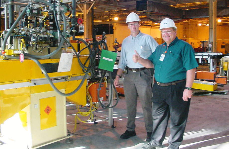 Don Fenton and another person posing and wearing hard hats in a factory with yellow machinery around them