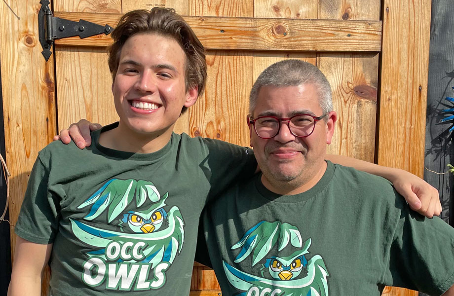 Cosmo Bray posing for a photo outdoors in front of a wooden door with his arm around his dad. they're both wearing the same green o c c owls t shirt
