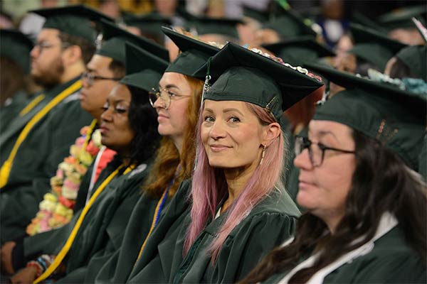 O C C students sitting at their graduation ceremony. One student is smiling at the camera, while the rest are watching the stage off frame.